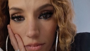 Close-up portrait of a woman with curly blonde hair resting her cheek on her hand, wearing dark eye makeup.