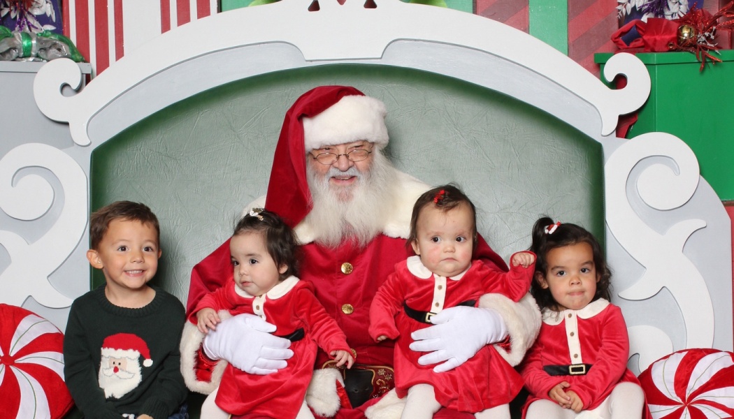 Santa Claus smiling on a festive chair, surrounded by four young children dressed in holiday outfits.