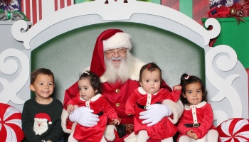 Santa Claus smiling on a festive chair, surrounded by four young children dressed in holiday outfits.