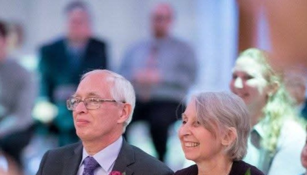 An older man with glasses and a suit sits next to a smiling grey-haired woman in an audience.