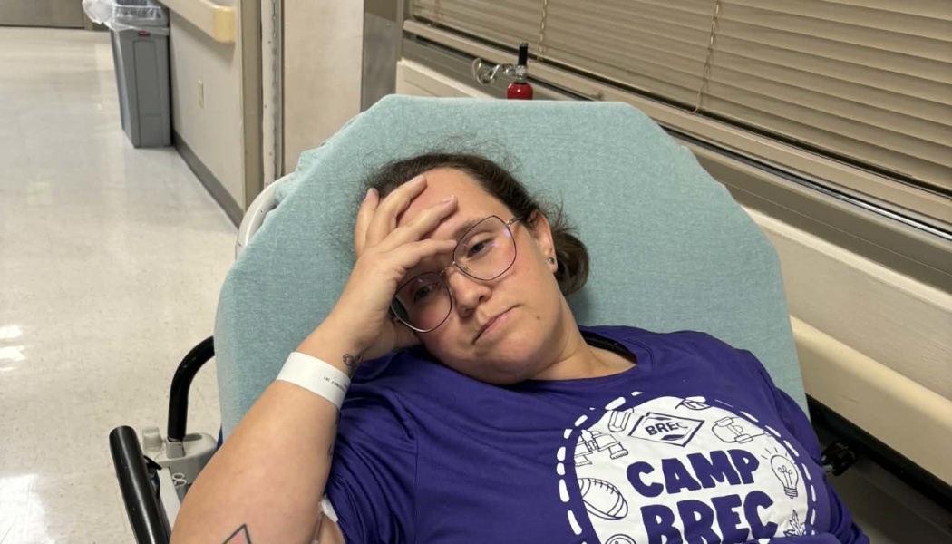 A tired woman wearing glasses lies on a hospital gurney with her hand resting on her forehead.