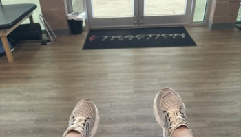 First-person view of feet in white running shoes facing the entrance doors of a sports facility.