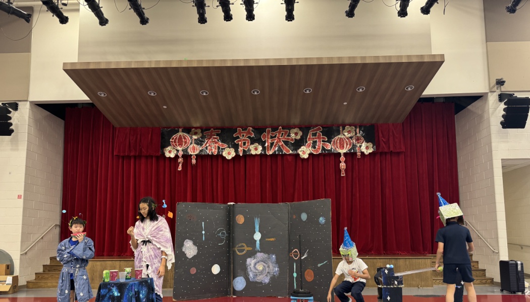Children in costumes perform a space-themed play in front of a stage decorated with a Chinese New Year banner.
