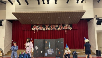 Children in costumes perform a space-themed play in front of a stage decorated with a Chinese New Year banner.