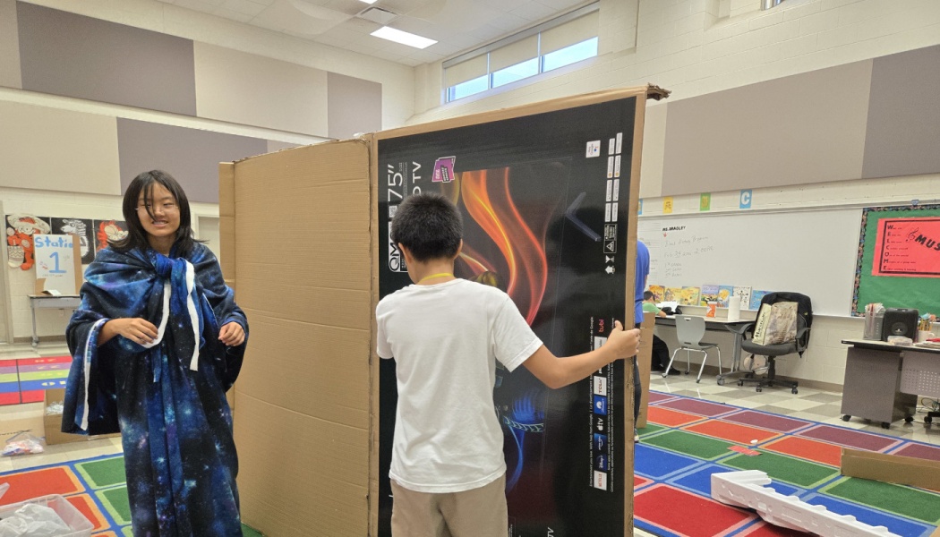 A boy holds up a large cardboard TV box while a girl wrapped in a galaxy blanket stands nearby in a classroom.