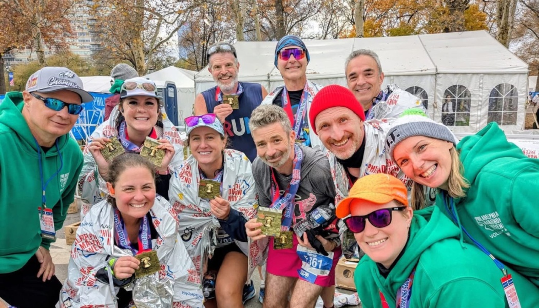 A group of smiling marathon runners wrapped in foil blankets and supporters display their medals together after a race.
