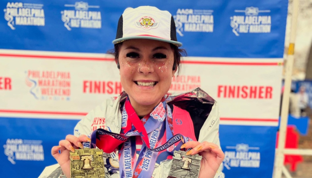 Smiling woman with glitter on her cheeks holds up two Philadelphia Marathon finisher medals.