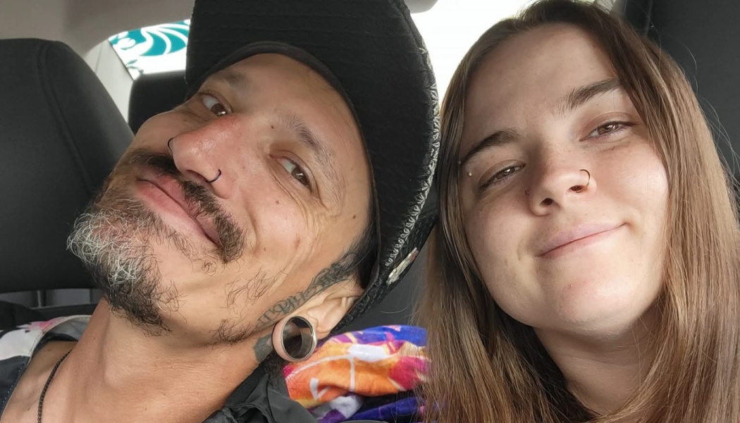 A smiling man with facial piercings and a cap poses close to a woman for a car selfie.