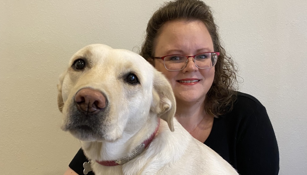 Smiling woman with red glasses posing closely behind her yellow Labrador retriever.