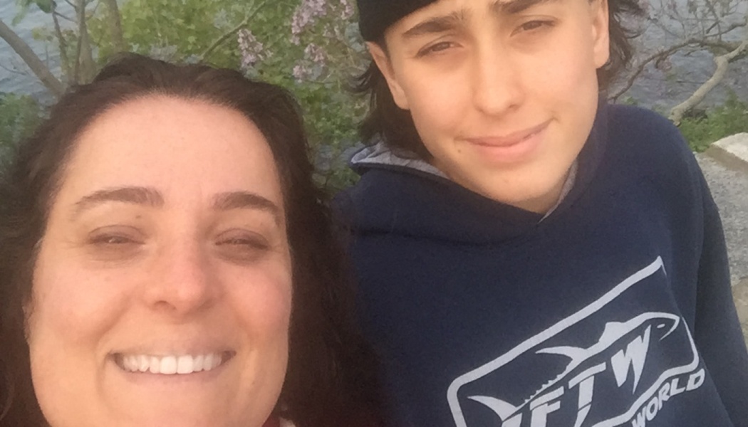 A smiling woman and a young man wearing a beanie and blue hoodie taking a selfie outdoors.
