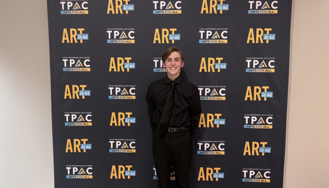 A smiling young man in formal black attire stands in front of a Tulsa Performing Arts Center backdrop.