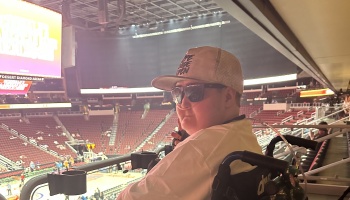 A person in a wheelchair wearing a cap, sunglasses, and a nasal cannula inside a large stadium arena.