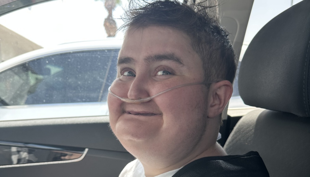 A young person wearing a nasal cannula smiles happily while sitting inside a car.