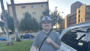 Smiling young person wearing a baseball cap and nasal cannula giving a thumbs-up outdoors.