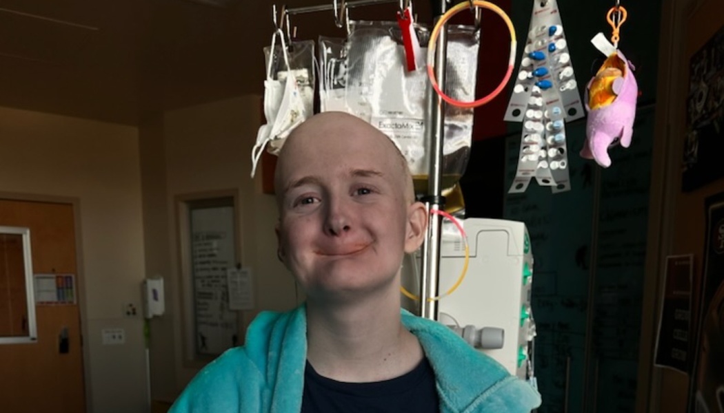 A smiling, bald young person wearing a turquoise hoodie in a hospital room in front of an IV pole.