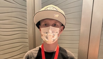 Young boy wearing a baseball cap, Disney cartoon face mask, and red lanyard looking at the camera.