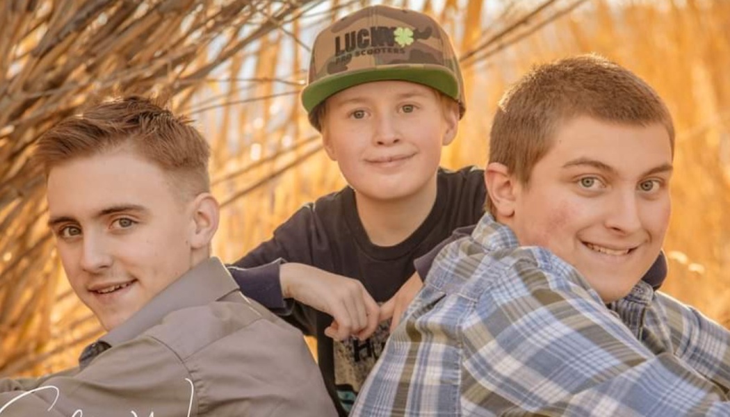 Three smiling boys, one wearing a camo cap, pose together outdoors in golden sunlight.