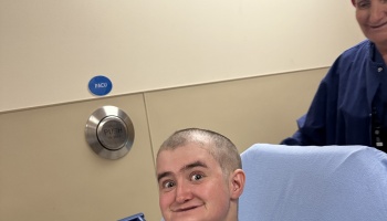 A smiling young person with a shaved head sits in a hospital bed in the PACU with a nurse nearby.