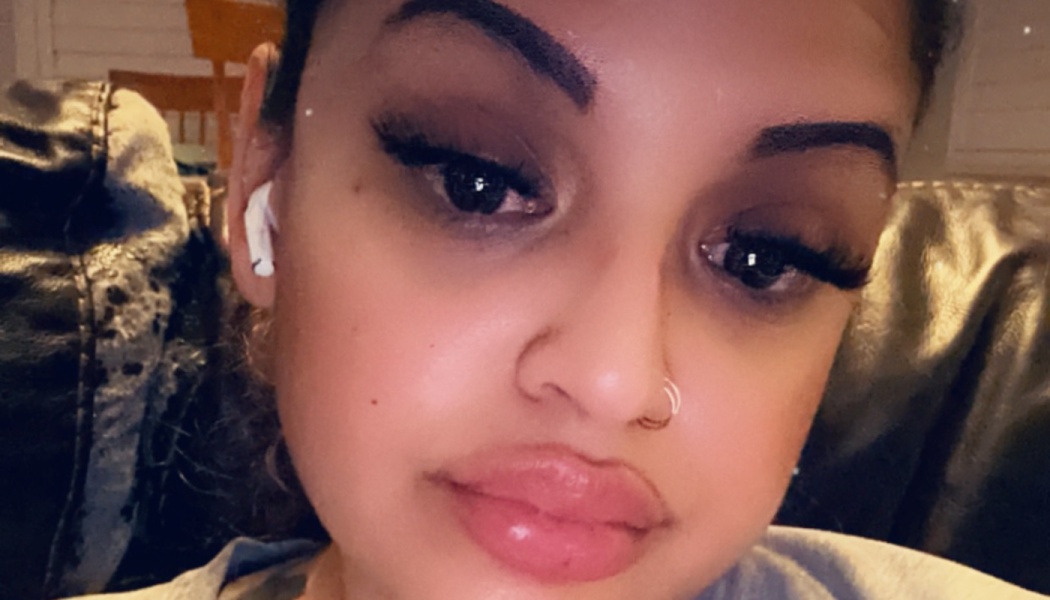 Close-up selfie of a woman with dark eyebrows, long eyelashes, a nose piercing, and a white earbud.