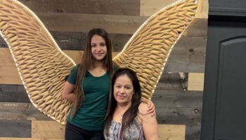 A young girl and a woman pose together in front of a wooden wall featuring large illuminated angel wings.