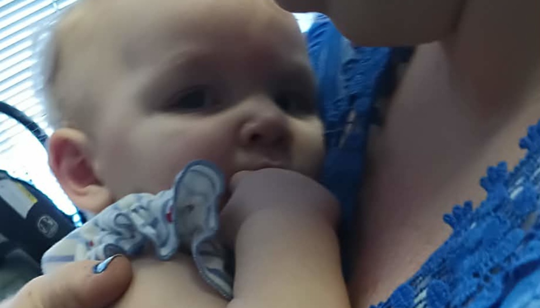 Close-up of a baby looking at the camera with their hand in their mouth while held by an adult.