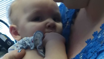 Close-up of a baby looking at the camera with their hand in their mouth while held by an adult.