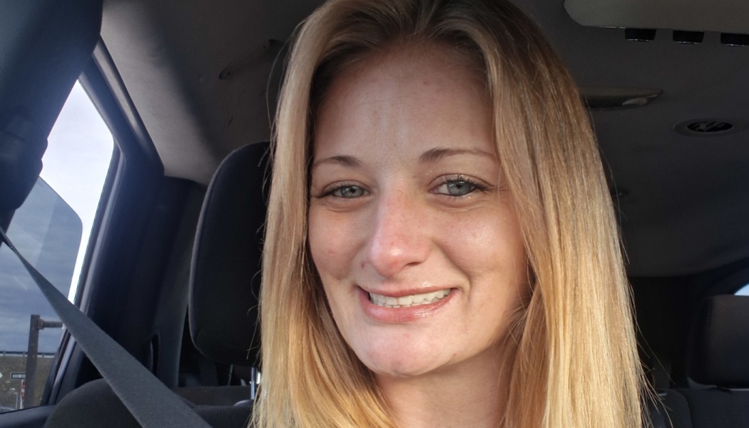 A smiling blonde woman sitting inside a car with her seatbelt on.