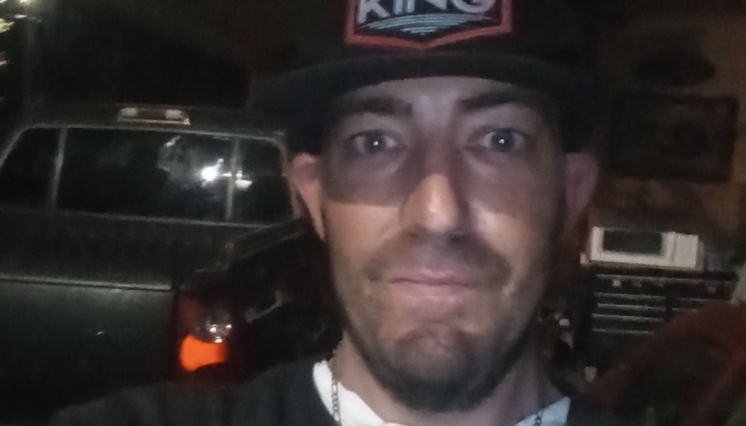 A man wearing a black baseball cap posing for a close-up selfie with a pickup truck behind him.