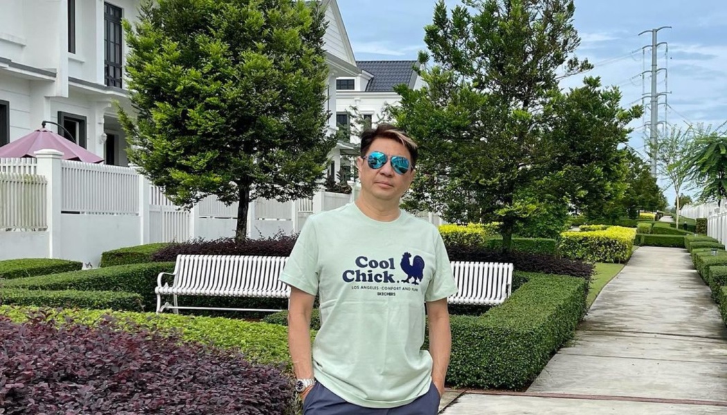 A man wearing sunglasses and a "Cool Chick" t-shirt stands outdoors in a landscaped neighborhood.