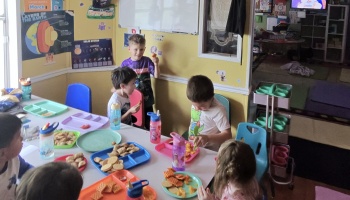 Young children gathered around a table enjoying snacks from colorful trays in a classroom.