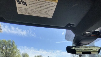 Interior view looking out a car windshield at a bright blue sky, showing the sun visor and rearview mirror.