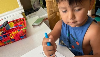 Young boy in a blue tank top intently colors on paper with a blue marker at a classroom table.