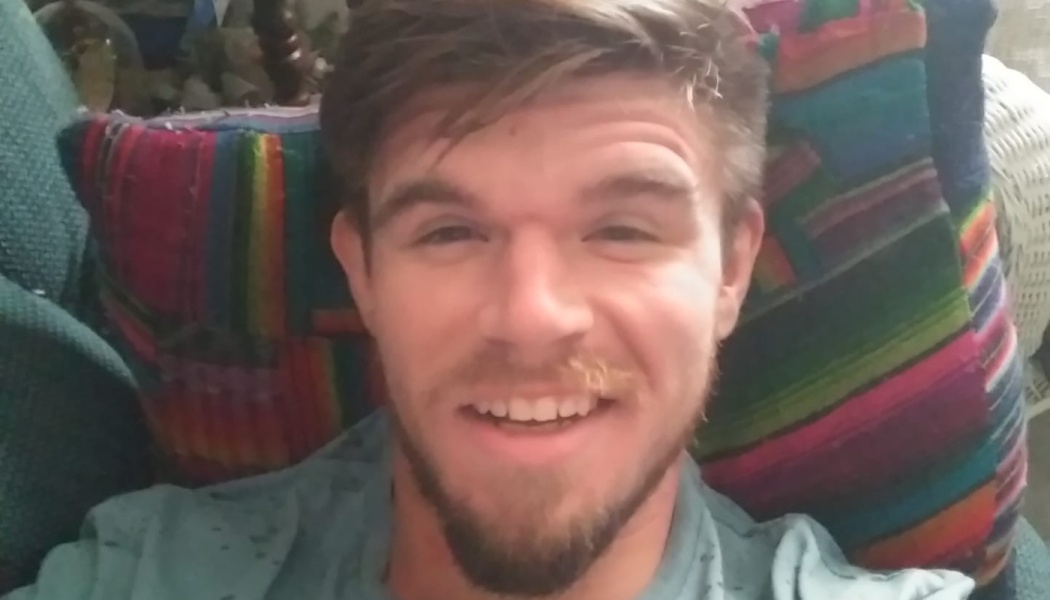 A smiling young man with a beard leaning back against a colorful patterned pillow.