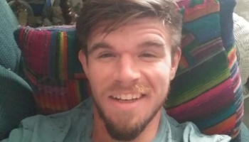 A smiling young man with a beard leaning back against a colorful patterned pillow.