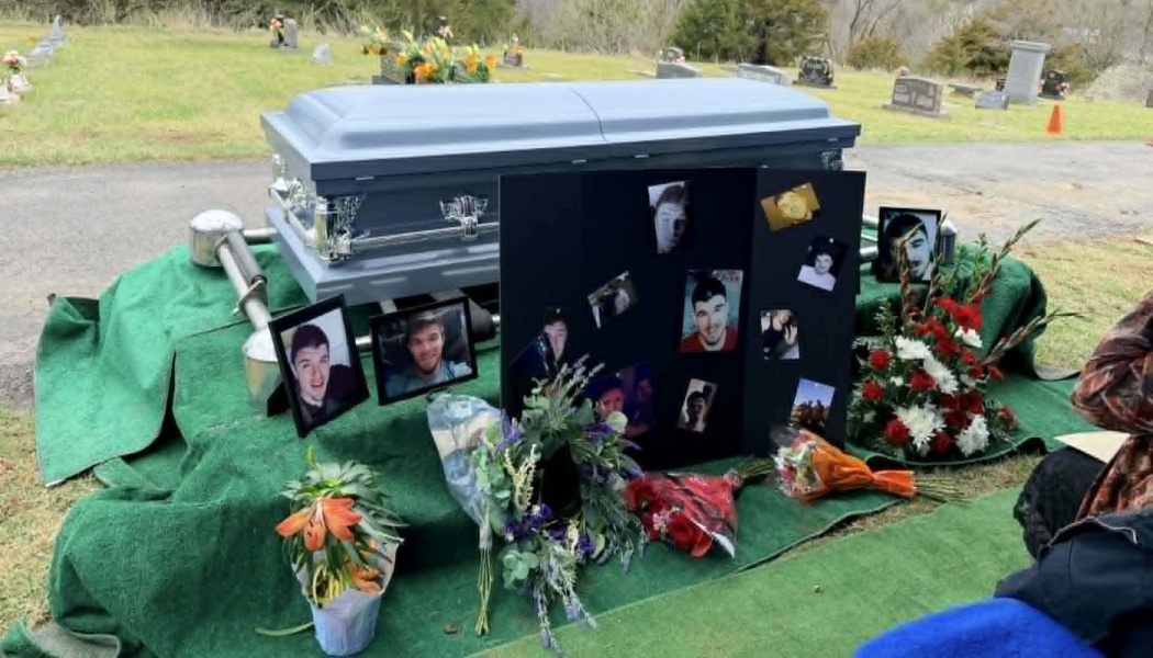 A light blue casket at a gravesite surrounded by flower bouquets and framed photos of a young man.