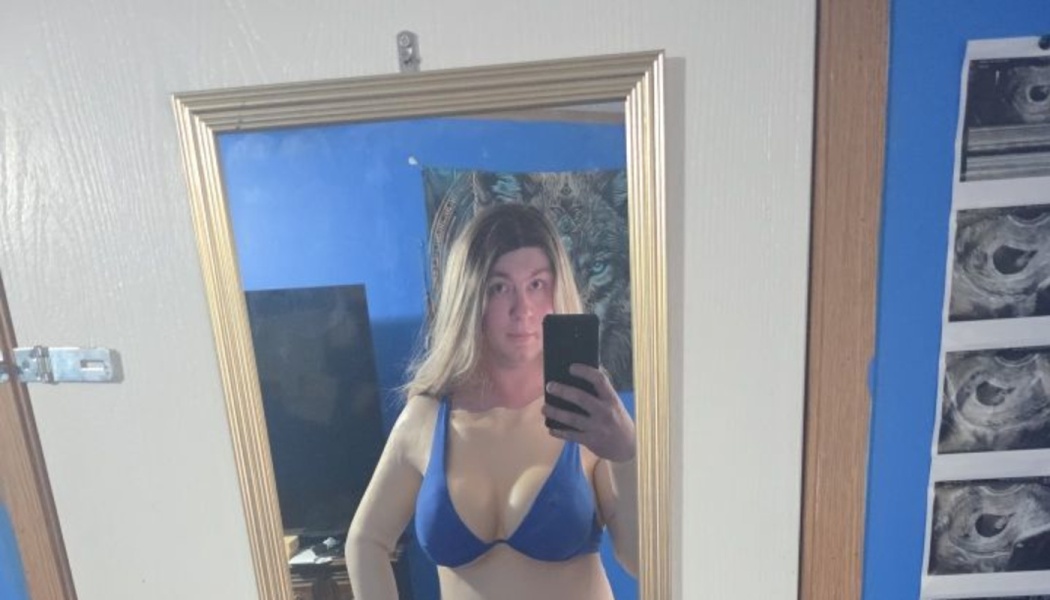 A person with long blonde hair taking a mirror selfie with a smartphone, wearing a blue bra top.