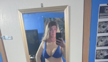 A person with long blonde hair taking a mirror selfie with a smartphone, wearing a blue bra top.