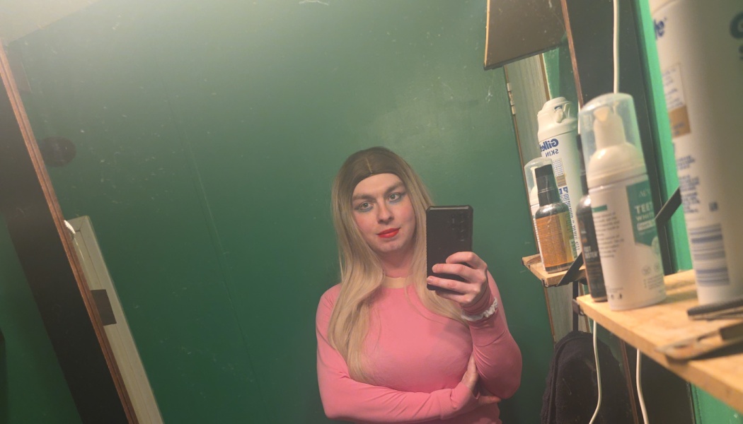 Blonde person with makeup wearing a pink top taking a mirror selfie in front of a green wall.