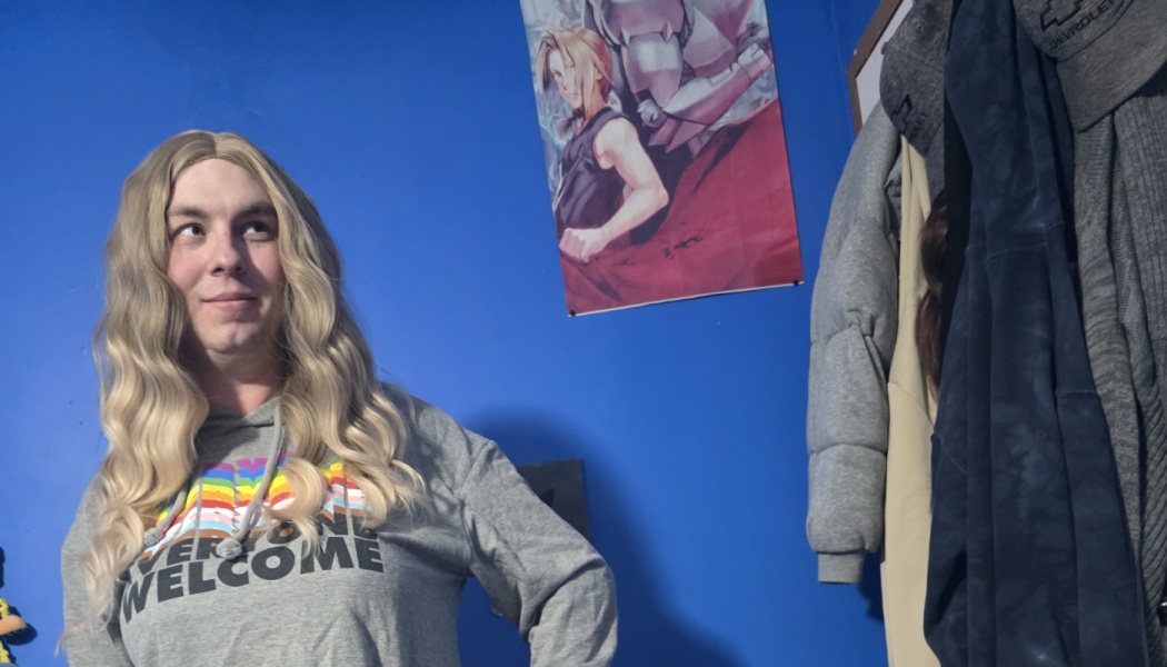 Person with long blonde hair wearing a grey "EVERYONE WELCOME" hoodie, smiling in front of a blue wall.