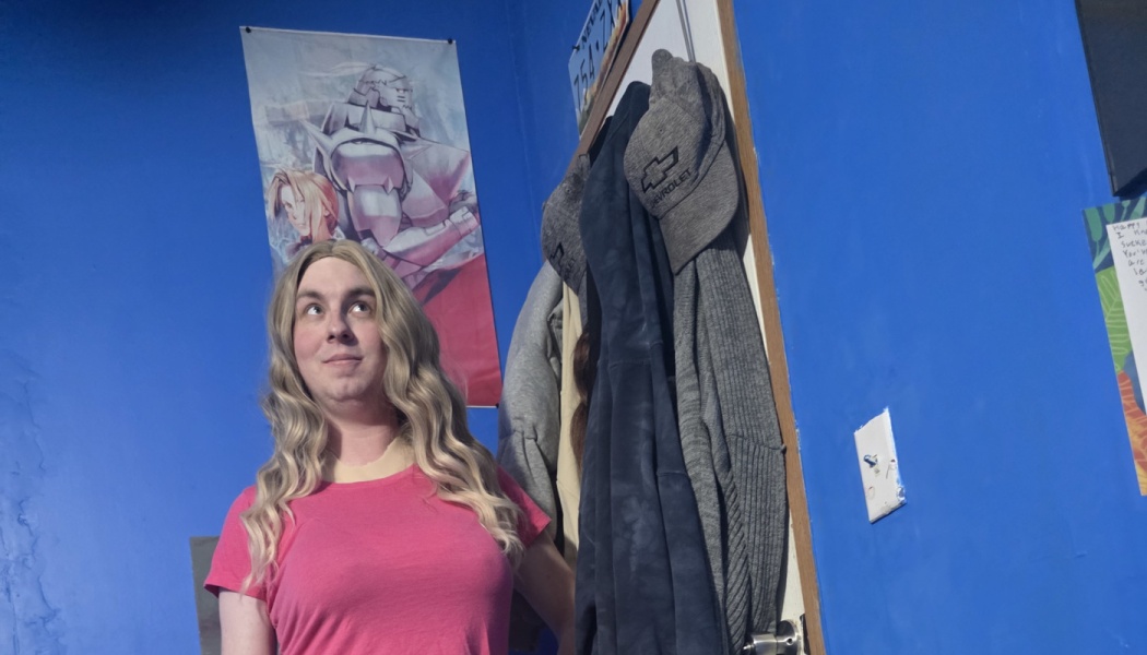 Person with wavy blonde hair and a pink shirt looking upward in a bright blue room with an anime wall poster.