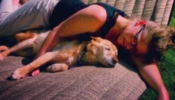 A woman with glasses resting outdoors on a cushion, hugging her sleeping tan dog closely.