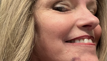 Close-up profile of a blonde woman smiling, wearing eye makeup and red lip liner.