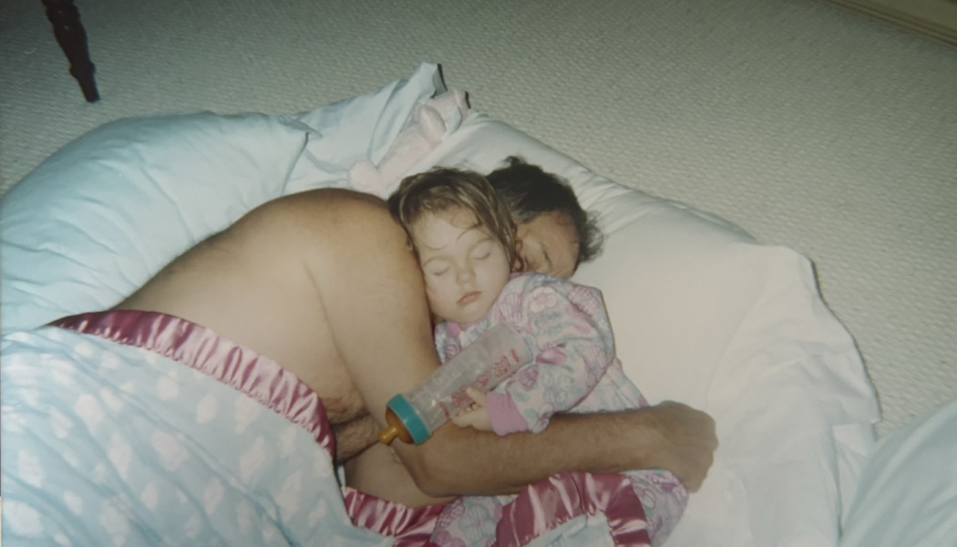 A sleeping toddler in pink pajamas holding a baby bottle cuddles tightly with a man on white pillows.