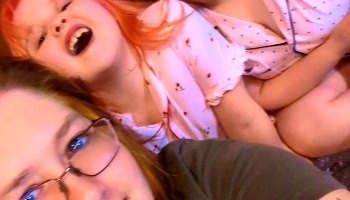 Woman with glasses lying next to a laughing red-haired child wearing pink pajamas.