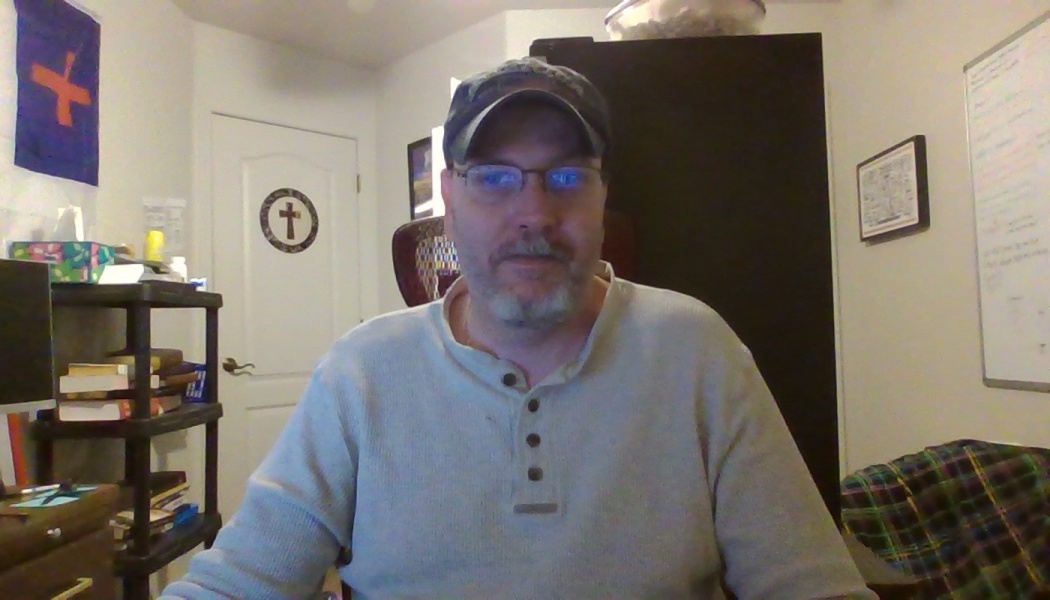 Middle-aged man with glasses, a beard, and a baseball cap wearing a gray henley shirt indoors.