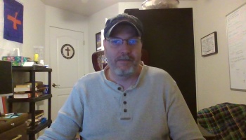 Middle-aged man with glasses, a beard, and a baseball cap wearing a gray henley shirt indoors.