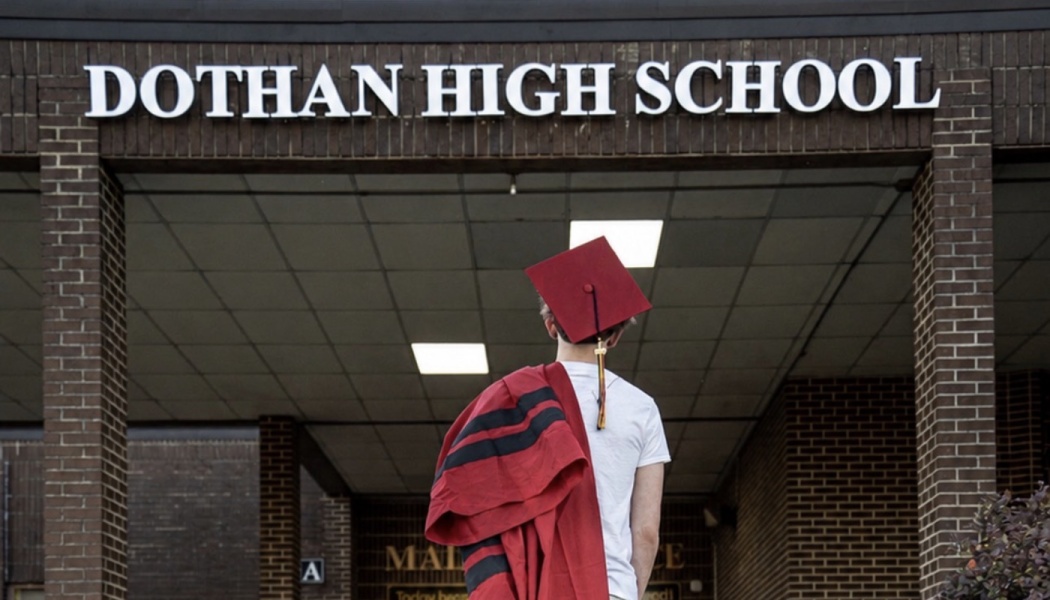 A student in a red graduation cap holds their gown over their shoulder, looking up at the Dothan High School sign.