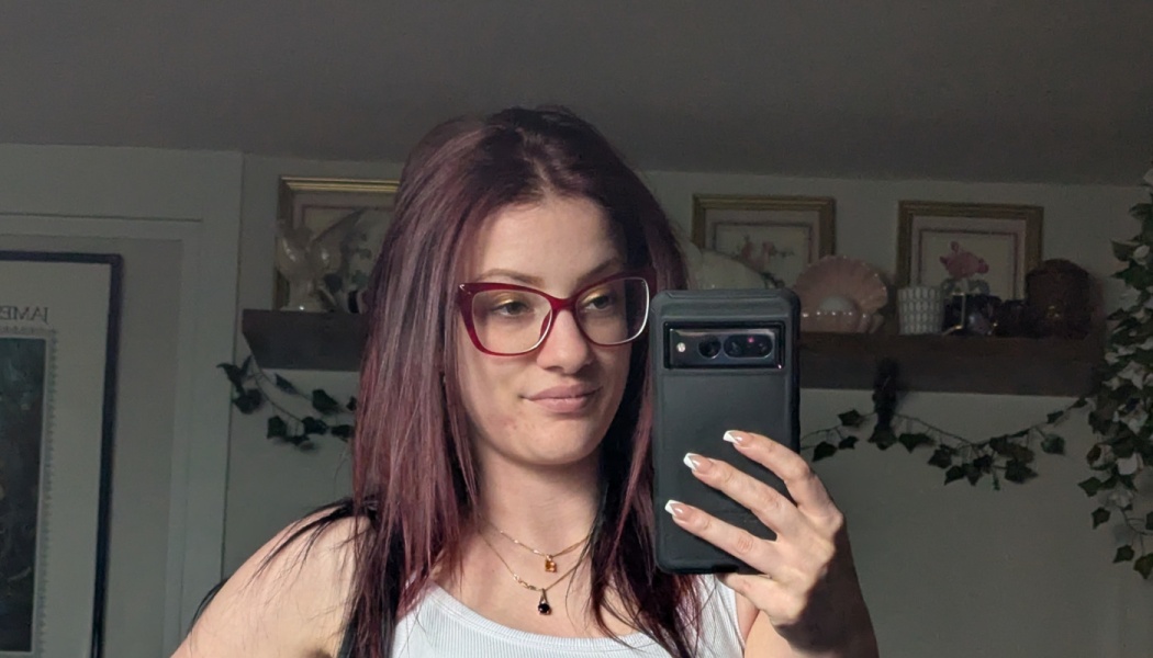 Woman with burgundy hair and red glasses taking a mirror selfie with her smartphone.