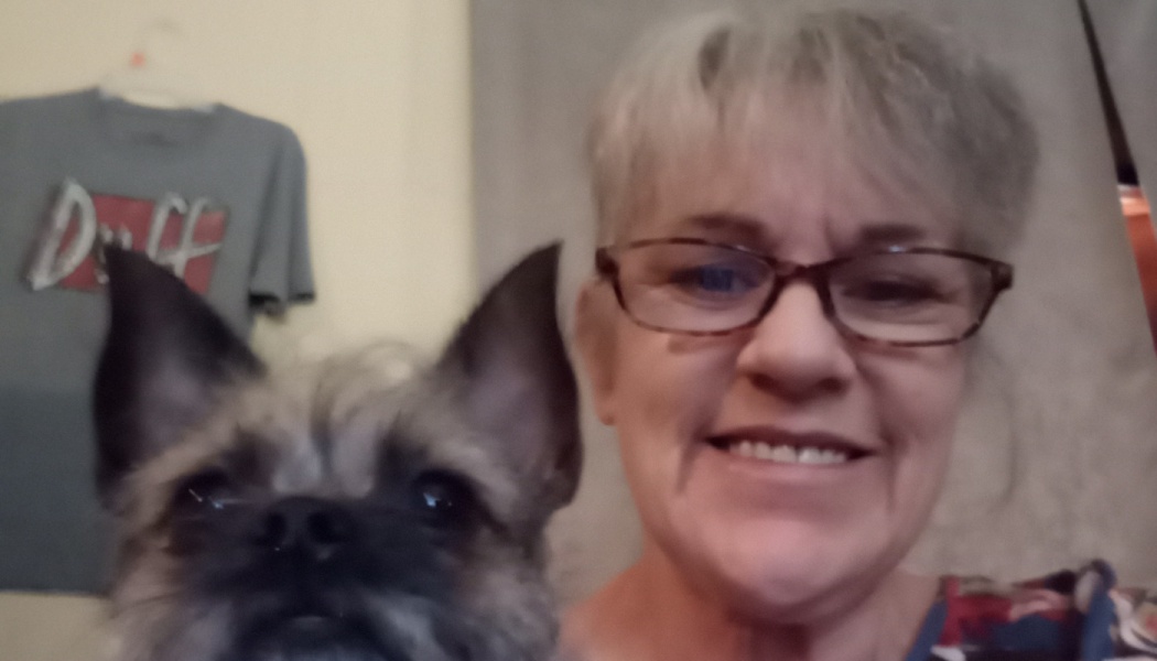 Smiling woman with short grey hair and glasses posing close to her small, scruffy grey dog.