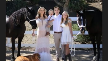 A smiling family dressed in white poses outdoors, flanked by two dark horses and a golden dog at their feet.